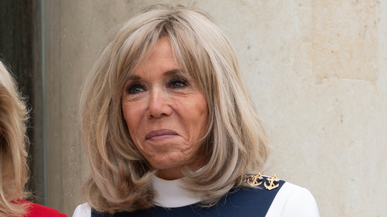 Brigitte Macron smirking while wearing a white dress at Elysee palace.