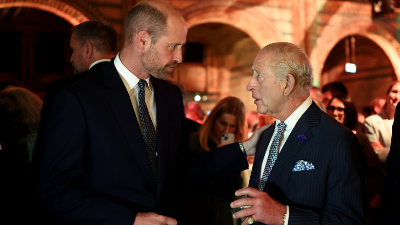 Prince William standing with his hand on King Charles' shoulder while they chat in suits