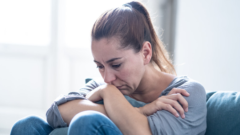 Woman with hair in ponytail sitting on a couch with her arms crossed and legs bent looking down with a worried or sad look in her face