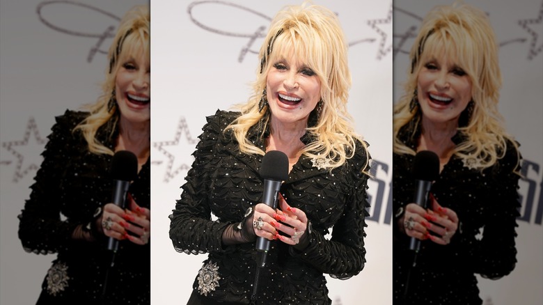 Dolly Parton wears a textured black dress and smiles while holding a microphone.