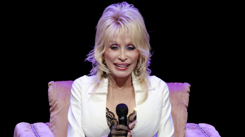 Dolly Parton wears colorful eyeshadow and a white blazer while smiling and holding a microphone.