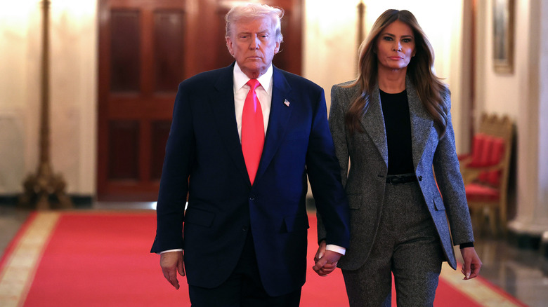 Donald and Melania Trump looking serious while holding hands and walking down a hallway