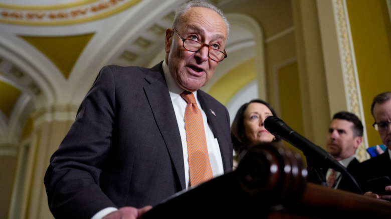 Chuck Schumer addressing an audience behind a podium in a dark suit with an orange tie