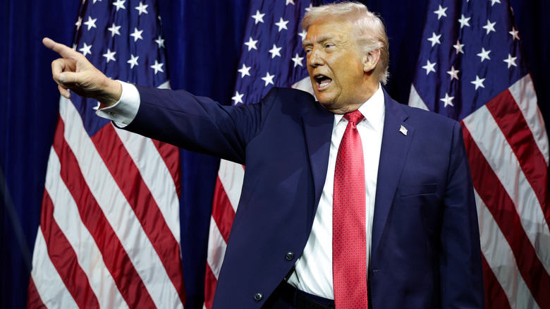Donald Trump pointing at someone while yelling in a navy suit and a red tie in front of American flags