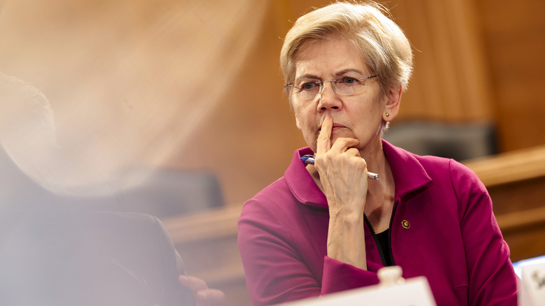 Elizabeth Warren looking pensive with a finger to her face in glasses and a purple top