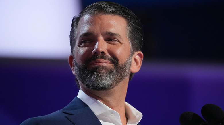 Donald Trump Jr. grinning in a navy suit in front of a microphone