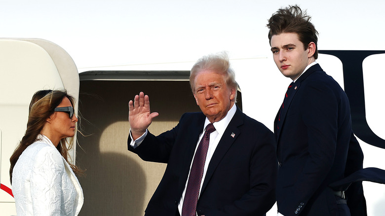 Donald Trump smirking and waving his hand while wearing a suit alongside Barron Trump and Melania Trump.