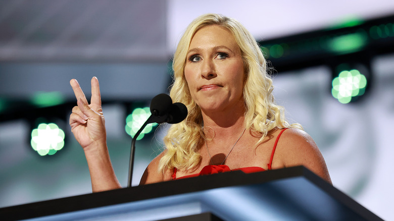 Marjorie Taylor Greene holding up a peace sign in a strappy red dress in front of a microphone
