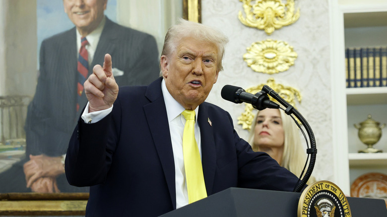 Donald Trump mid talk, hand raised, behind a podium in the Oval Office.
