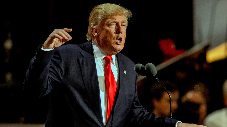 Donald Trump at a podium in a suit with his hand raised in a gesture as he speaks.