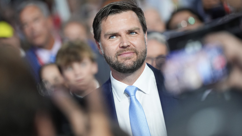 JD Vance smirking in a suit with a blue tie surrounded by people