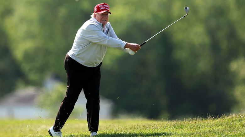 Donald Trump taking a swing on the golf course in a red MAGA hat while gurning
