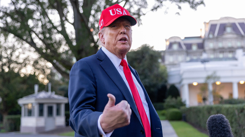 Donald Trump gesturing with his thumb in a blue suit with a red tie and red USA hat