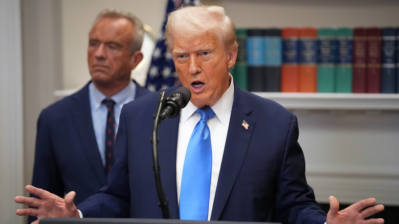 Donald Trump giving a speech while raising his hands in a navy suit with a light blue tie