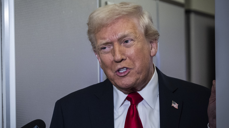 Donald Trump speaking on Air Force One in a black suit with a red tie