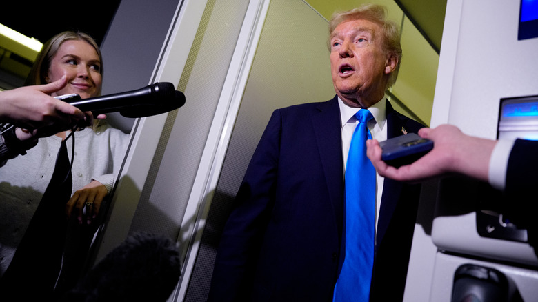 Donald Trump addressing reporters on Air Force One while Karoline Leavitt looks on smiling proudly