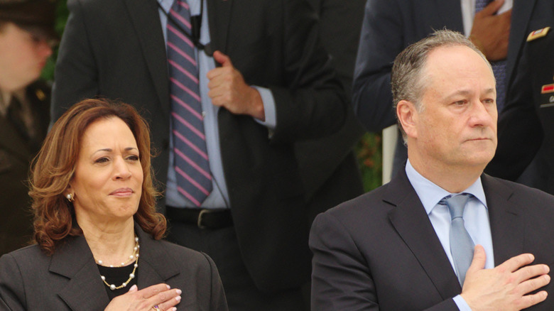 Doug Emhoff in a suit standing next to Kamala Harris who's wearing a black blazer and shirt while standing for the national anthem.