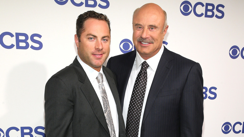 Dr. Phil and Jay McGraw wearing suits on the red carpet with a CBS logo in the background in 2016