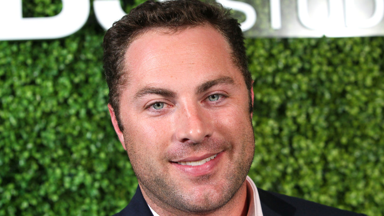 Dr. Phil's son Jay McGraw smiles in front of a green background in a close-up shot on the red carpet