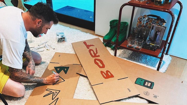 Jordan McGraw sits on the ground writing the name of his band, BORT, on two cardboard signs.