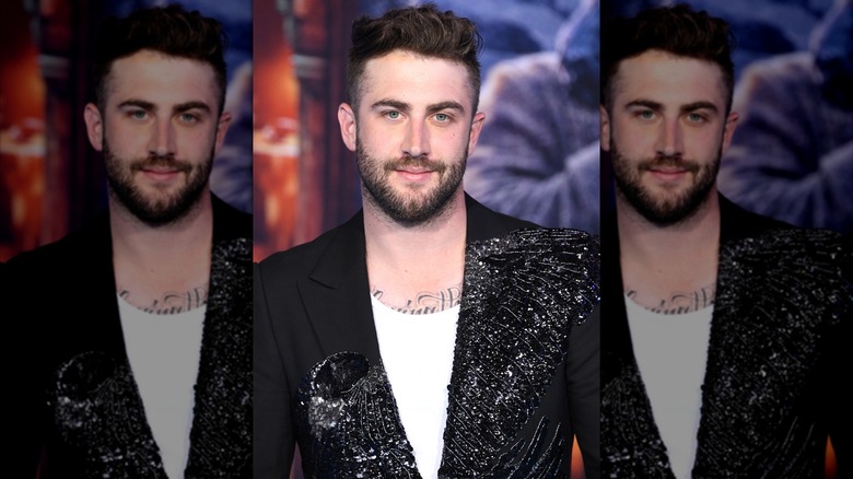 Dr. Phil's son Jordan McGraw smiles wearing a sequin black suit with a white tank top underneath that reveals a chest tattoo.