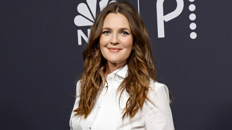 Drew Barrymore smiling and posing wearing a white collared top, and her long brown hair down.