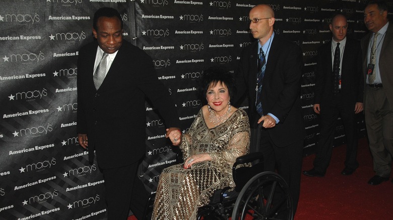 Jason Winters holding Elizabeth Taylor's hand as another man wheels her in a wheelchair