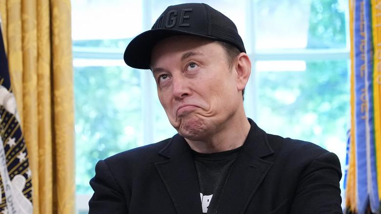 Elon Musk wearing a black DOGE hat and frowning while in the Oval Office