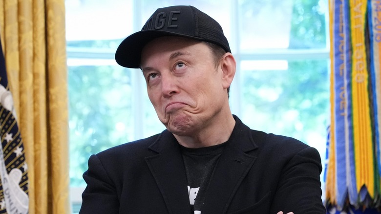 Elon Musk in all-black outfit in the Oval Office making an exaggerated frown expression.