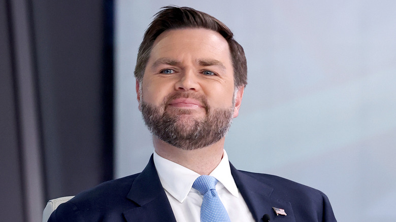 JD Vance smirking in a navy suit with a light blue tie and an American flag pin