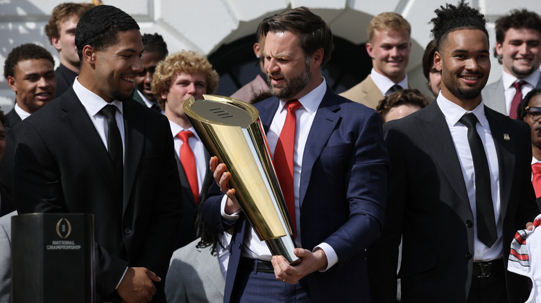 JD Vance looking utterly perplexed as he holds the top of the Ohio state championship trophy in a navy suit