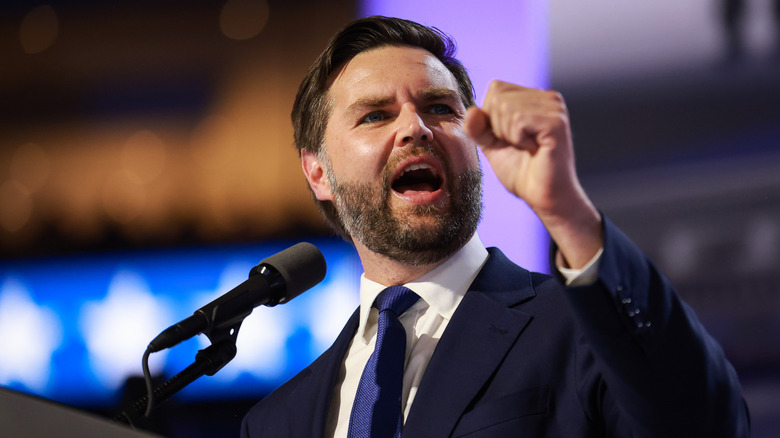 JD Vance animatedly shaking his fist in front of a microphone in a navy suit