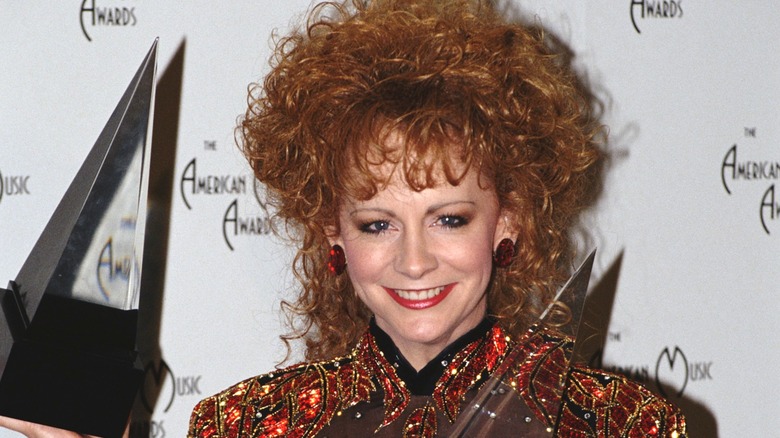 Reba McEntire holding awards, smiling, very big curly hair on top and hanging down