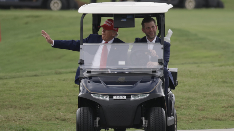 Eric Trump driving Donald Trump around in a golf cart while he waves to his fans