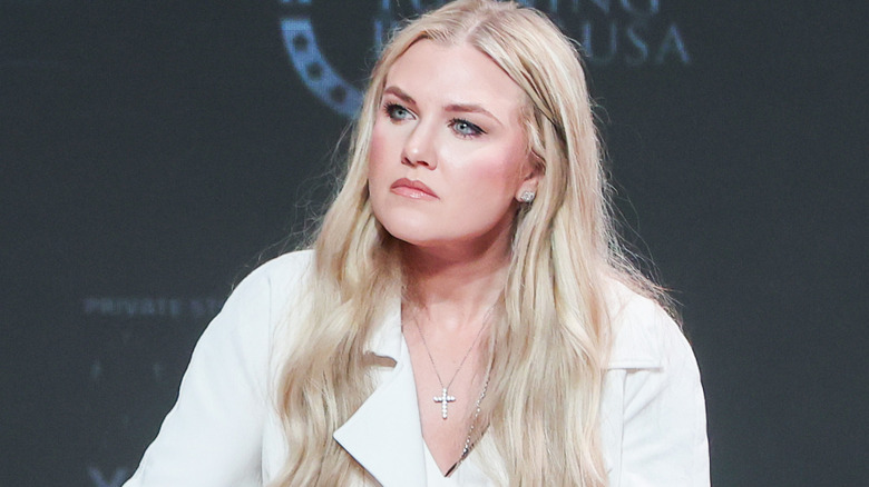 Erika Kirk looking very serious in a white blazer, white top, and a sparkly cross necklace