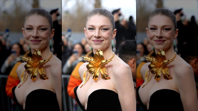 Hunter Schafer posing in a black dress with a giant gilded flower around her neck