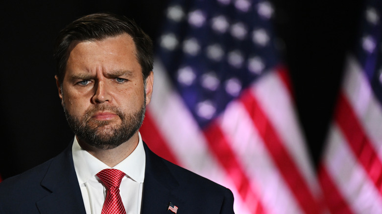 JD Vance looking glum in a suit in front of an American flag