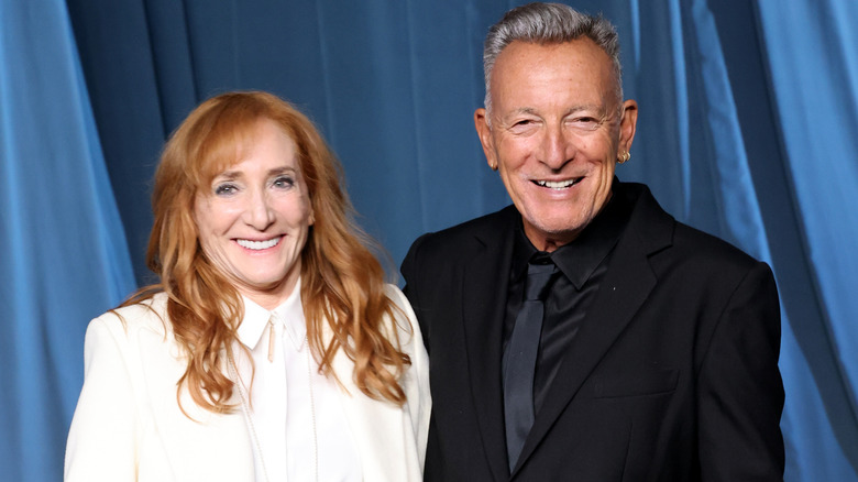 Patti Scialfa and Bruce Springsteen smiling while posing together in front of a light blue curtain