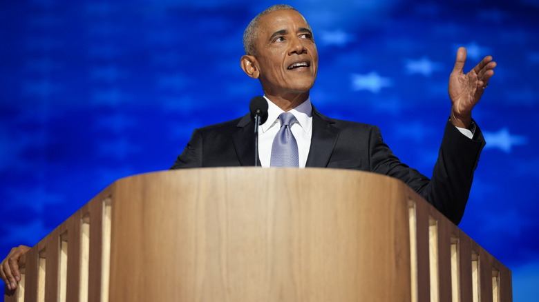 Barack Obama giving a speech in a dark suit in front of a blue background
