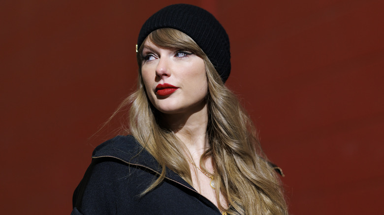 Taylor Swift looking over her shoulder in a red hall, wearing a black beanie.