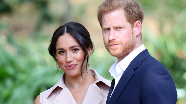 A smiling Meghan Markle standing next to a serious-looking Prince Harry