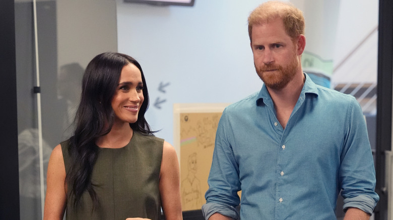A smiling Meghan Markle standing next to a serious-looking Prince Harry
