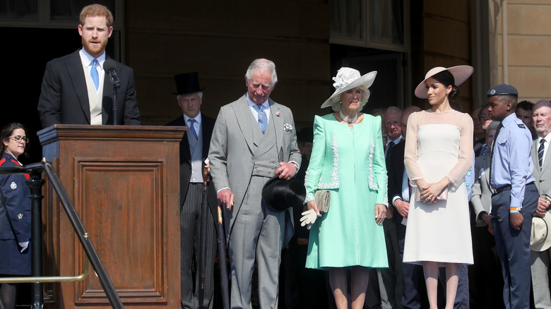 Prince Harry delivering a speech while King Charles III, Queen Camilla, and Meghan Markle look on