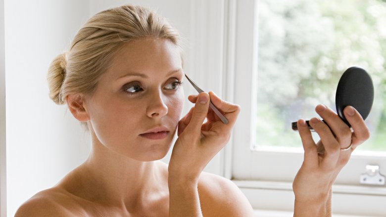 woman plucking eyebrows while looking in compact mirror