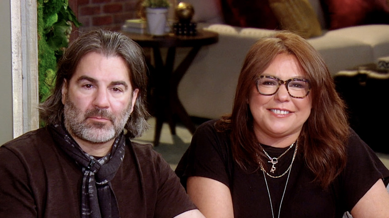 Rachael Ray smiling in a black shirt while wearing glasses next to John Cusimano who's wearing a brown t-shirt and scarf.