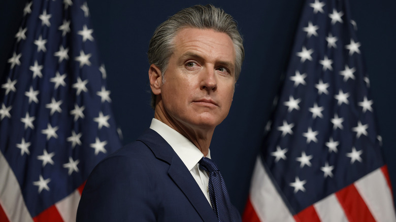 Gavin Newsom looking pensive in a suit in front of two American flags