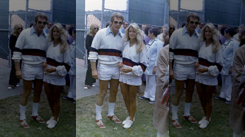 Lee Majors and Farrah Fawcett posing together in matching blue and red striped sweaters with dress shirts underneath and short white bottoms