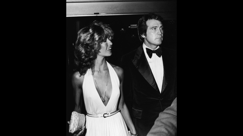 Farrah Fawcett and Lee Majors walking hand in hand, with Fawcett carrying a clutch and Majors in a black suit and tie