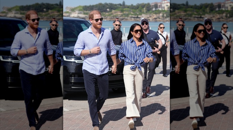 Prince Harry holding Meghan Markle's hand, the latter wearing sunglasses and striped shirt with beige pants.