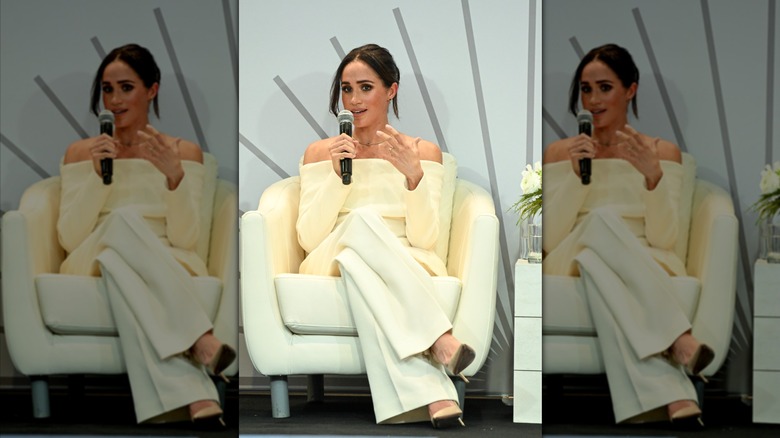 Meghan Markle wearing a cream off-the-shoulder shirt and white pants while speaking at a panel held by Project Healthy Minds.